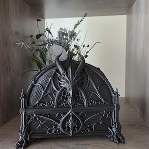 Könnte beinhalten: Eine schwarze dekorative Box mit einem Drachendesign. Die Box hat einen Drachenkopf auf dem Deckel, komplizierte Muster und Krallenfüße. Die Box wird auf einem Holzregal ausgestellt, mit einem Blumenstrauß im Hintergrund.