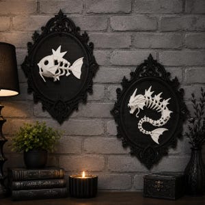 May include: Two oval black ornate frames containing white skeletal fish figures. The left frame features a small fish skeleton, while the right frame displays a larger, serpentine dragon-like fish skeleton. Both are mounted on a gray brick wall.