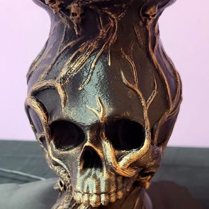Pode incluir: Um vaso preto e dourado em forma de caveira com um design detalhado. O vaso apresenta um rosto de caveira e é adornado com detalhes dourados, incluindo um padrão dourado em forma de videira. O vaso foi projetado para conter flores, com flores vermelhas visíveis no topo.