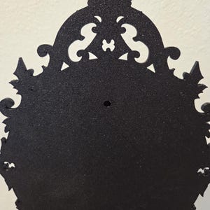 May include: A black, oval-shaped wall-mounted frame with a textured, matte finish. The frame features an ornate, decorative border with intricate scrollwork and pointed edges. A small, circular mounting hole is centered near the top section.