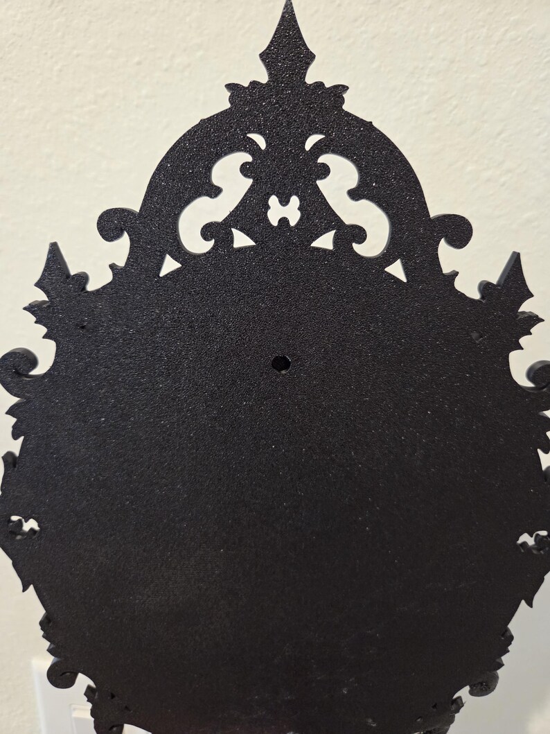 May include: A black, oval-shaped wall-mounted frame with a textured, matte finish. The frame features an ornate, decorative border with intricate scrollwork and pointed edges. A small, circular mounting hole is centered near the top section.