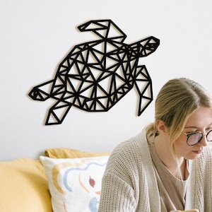 Könnte beinhalten: Eine Frau sitzt auf einem Bett mit einem Laptop und einem Telefon in den Händen. An der Wand hinter ihr befindet sich eine geometrische schwarze Schildkröten-Wanddekoration.