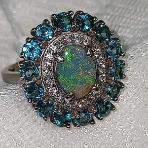 May include: A silver ring with an oval opal gemstone displaying green and gold iridescence. The opal is encircled by a halo of small, clear gemstones and a border of blue gemstones, forming a floral pattern. The ring is set on a silver band.