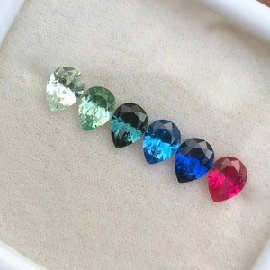 Può includere: Una fila di sei pietre preziose a forma di pera in una scatola di presentazione bianca. Le pietre preziose sono disposte da sinistra a destra in un gradiente di colori: verde chiaro, verde, verde-azzurro, turchese, blu e rosso.