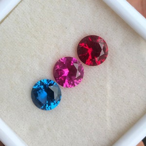 May include: Three round gemstones in a white display box. The gemstones are blue, pink, and red.