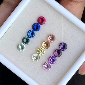 May include: A collection of ten round gemstones in a white display box. The gemstones are arranged in a rainbow pattern, starting with red and ending with purple.