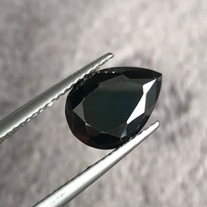 May include: A black pear-shaped gemstone held by tweezers. The gemstone is faceted and has a high polish.
