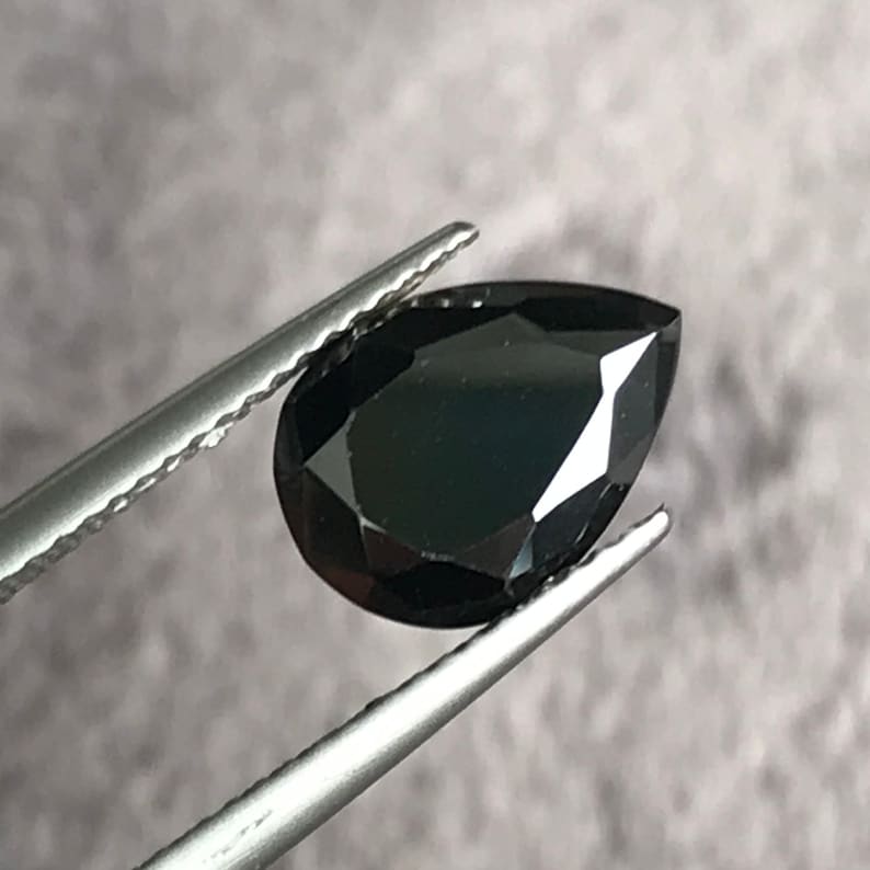 May include: A black pear-shaped gemstone held by tweezers. The gemstone is faceted and has a high polish.