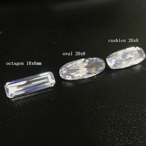 Könnte beinhalten: Vier klare Edelsteine in verschiedenen Formen und Größen auf einer schwarzen Oberfläche. Die Edelsteine sind mit "octagon 18x6mm", "oval 20x8" und "cushion 20x8" beschriftet.