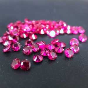 May include: A scattering of bright pink gemstones, likely rubies, on a dark grey surface. The gemstones are faceted and reflect light.