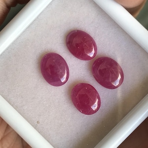May include: Four oval-shaped, polished ruby gemstones. The rubies are a deep, rich red color and are displayed in a white, rectangular box. The gemstones have a smooth, glossy surface.