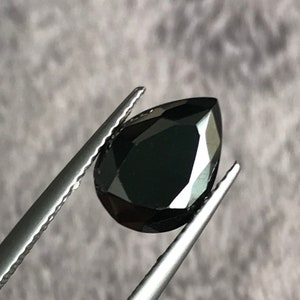 May include: A black pear-shaped gemstone held by tweezers. The gemstone is faceted and has a high polish.