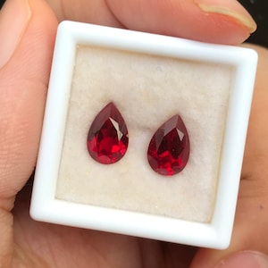 Op de afbeelding: Twee peervormige rode edelstenen in een witte doos. De edelstenen zijn gezet in een witte doos met een witte achtergrond.