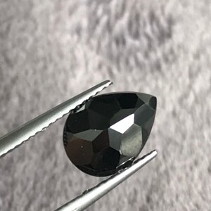 May include: A black pear-shaped gemstone with a faceted cut, held by tweezers.