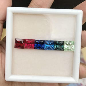 Peut inclure: Une boîte blanche contenant une rangée de sept pierres précieuses de forme carrée. Les pierres précieuses sont rouges, bleues et vertes.