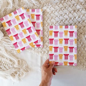 Może przedstawiać: Trzy białe karty z powtarzającym się wzorem różowych, żółtych i czerwonych kolumn z nadrukowanymi słowami "Doric", "Ionic" i "Corinthian".