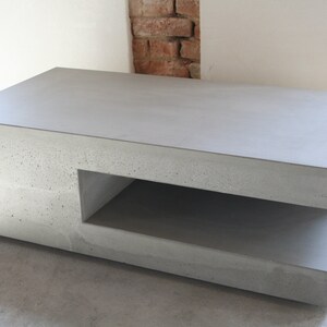 Könnte beinhalten: Ein grauer Beton-Couchtisch mit einem Regal darunter. Der Tisch hat eine raue, strukturierte Oberfläche.