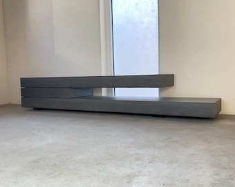 Puede incluir: Una consola multimedia rectangular de perfil bajo en color gris oscuro, con un diseño escalonado de dos niveles horizontales distintos. La estructura está compuesta por bloques sólidos de superficie lisa dispuestos en forma.