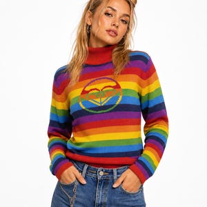 May include: A long-sleeve turtleneck jumper featuring horizontal rainbow stripes in red, orange, yellow, green, blue, and purple. A circular logo graphic is centred on the chest. The jumper is paired with blue denim jeans featuring a silver chain.