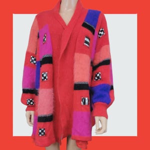 May include: A colourful, open-front cardigan in shades of red, pink, and purple. The sweater has a patchwork design with black and silver accents, and small square embellishments. The sleeves are long and the cardigan is knee-length.