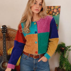 May include: A long-sleeve jacket made of multi-coloured suede panels in shades of red, blue, teal, purple, green, and tan. The jacket features a round neckline, a front button closure with four colourful buttons, and two small horizontal front pockets.