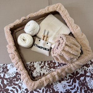 Op de afbeelding: Een beige, pluche frame met een wit canvas, twee bollen witte garen, twee houten naalden en een klein wit zakje met de tekst "ETHNOPUNCH" erop gedrukt.