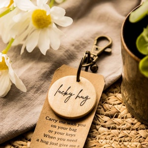 May include: A wooden keychain with a round charm that says "lucky hug". The charm is attached to a silver key ring. The keychain is sitting on a woven mat with a brown fabric in the background.