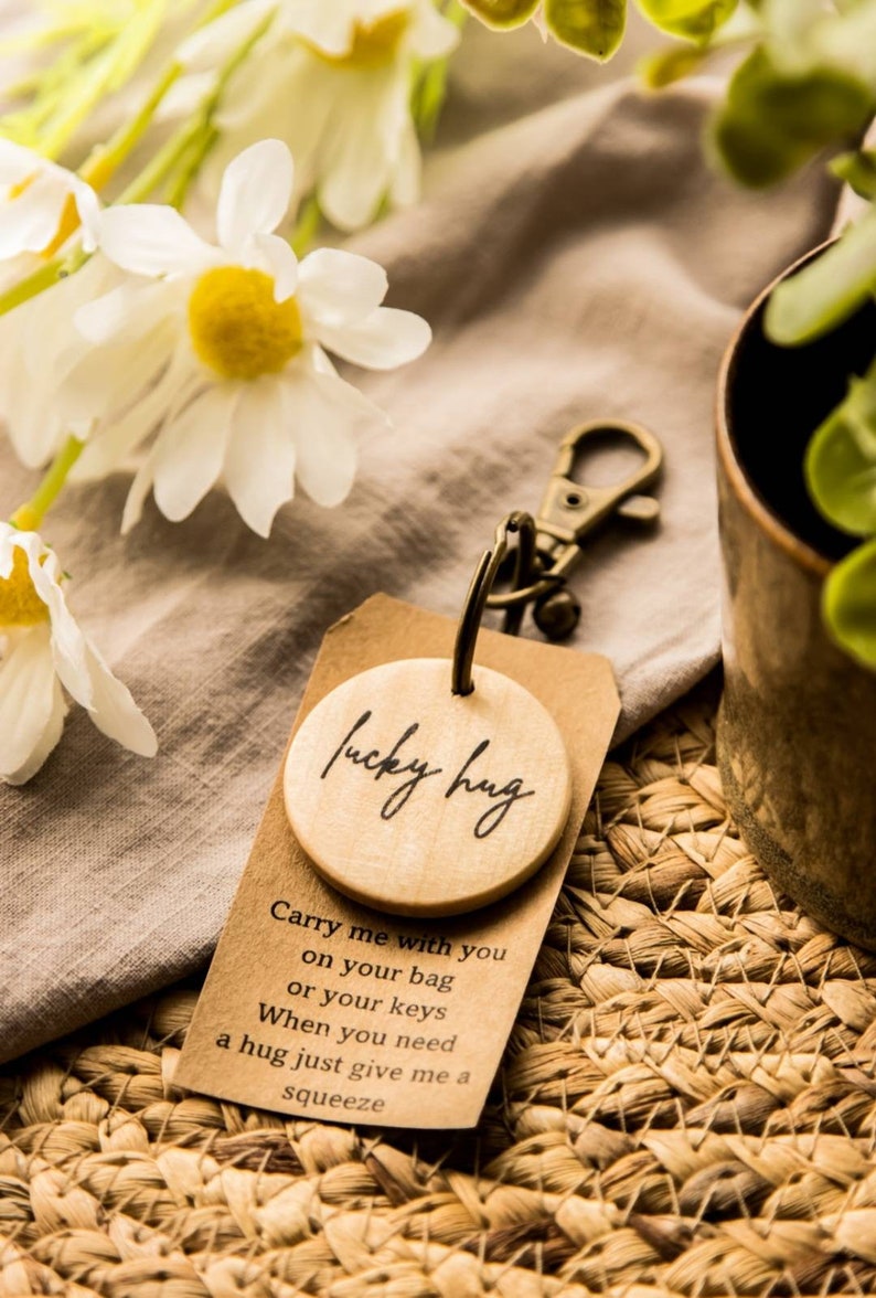 May include: A wooden keychain with a round charm that says "lucky hug". The charm is attached to a silver key ring. The keychain is sitting on a woven mat with a brown fabric in the background.