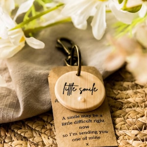 May include: A wooden keychain with a small round wooden charm that says "little smile". The charm has a small white pearl in the center. The keychain is accompanied by a small card with the text "A smile may be a little difficult right now so I'm sending you one of mine x".