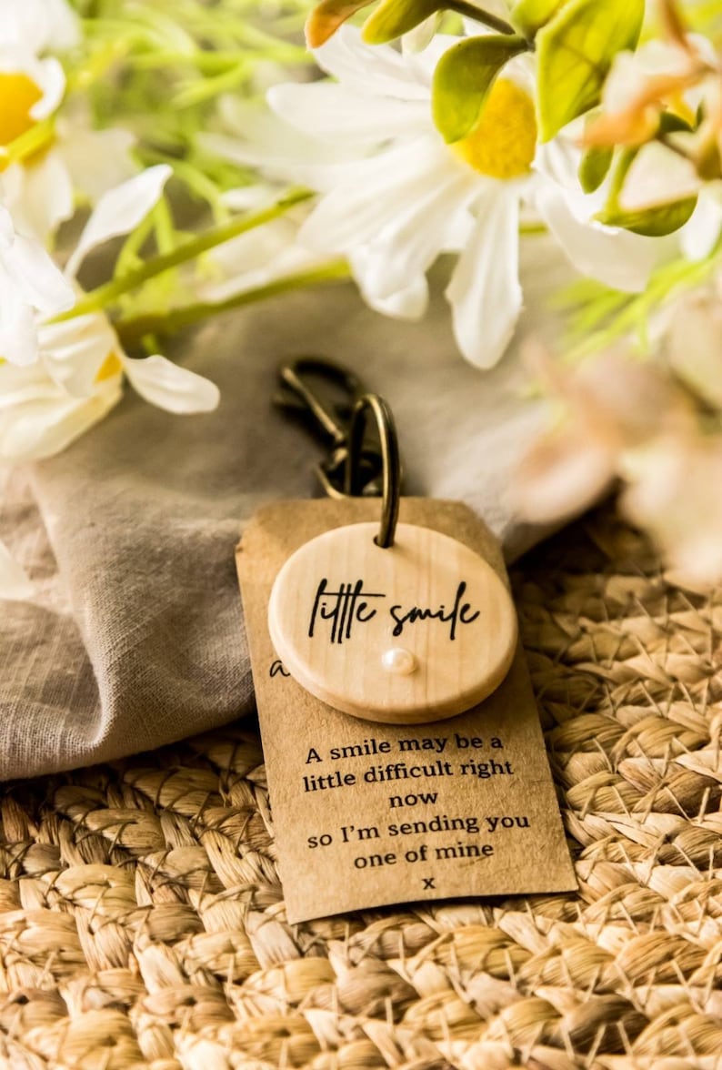 May include: A wooden keychain with a small round wooden charm that says "little smile". The charm has a small white pearl in the center. The keychain is accompanied by a small card with the text "A smile may be a little difficult right now so I'm sending you one of mine x".