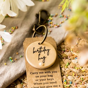 May include: A wooden keychain with a round tag that says "birthday hug" and a small pearl. The tag is attached to a metal key ring. The keychain is sitting on a brown woven surface with colorful sprinkles.
