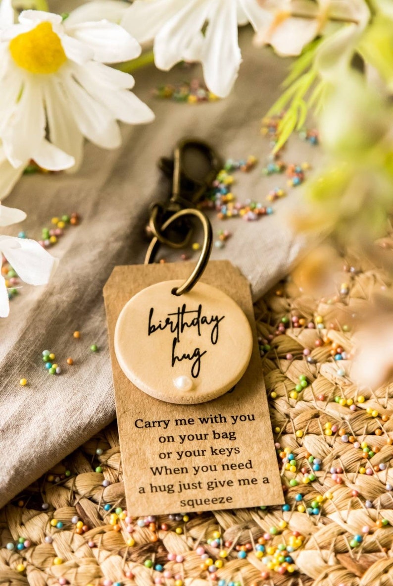 May include: A wooden keychain with a round tag that says "birthday hug" and a small pearl. The tag is attached to a metal key ring. The keychain is sitting on a brown woven surface with colorful sprinkles.