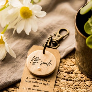 May include: A wooden keychain with a small round wooden tag that says "little smile" with a small white pearl in the center. The tag is attached to a brown paper tag with the text "A smile may be a little difficult right now so I'm sending you one of mine X".