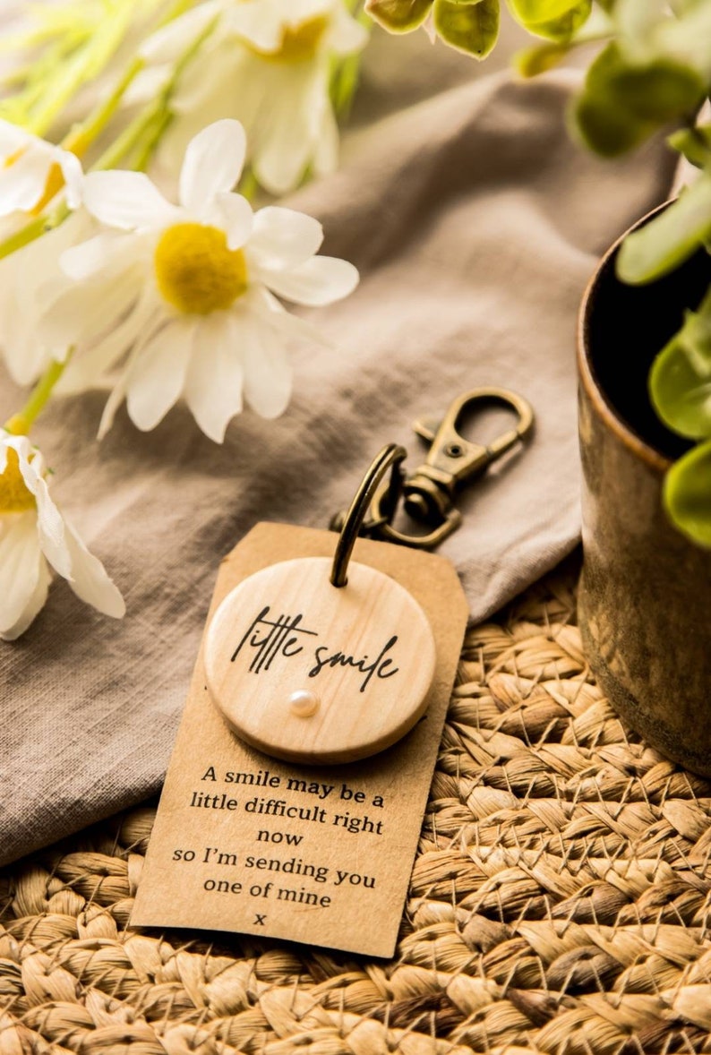 May include: A wooden keychain with a small round wooden tag that says "little smile" with a small white pearl in the center. The tag is attached to a brown paper tag with the text "A smile may be a little difficult right now so I'm sending you one of mine X".