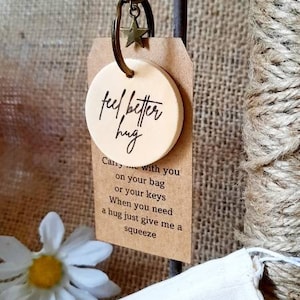 May include: A wooden keychain with a round tag that says "feel better hug". The tag is attached to a metal key ring with a star charm. The keychain is accompanied by a small fabric bag with the text "JUST FOR YOU THE little hug FAMILY" printed on it.
