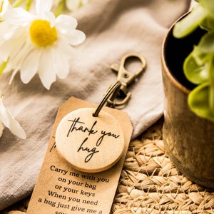 May include: A wooden keychain with a round wooden charm that says "Thank you hug". The charm is attached to a brown card with the text "Carry me with you on your bag or your keys. When you need a hug just give me a squeeze."