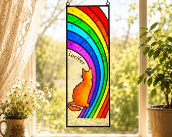 May include: A rectangular stained glass panel featuring a rainbow and an orange cat silhouette. The word "Lucifer" is written on the panel. The panel is suspended by a chain and hangs in front of a window.