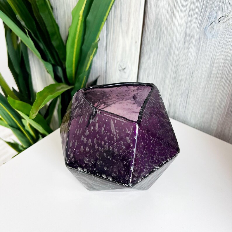 Puede incluir: Un jarr&oacute;n de cristal facetado de color amatista con un patr&oacute;n moteado. El dise&ntilde;o geom&eacute;trico presenta un borde oscuro y una amplia abertura. El jarr&oacute;n est&aacute; sobre una superficie blanca con follaje verde en el fondo, creando una est&eacute;tica moderna.