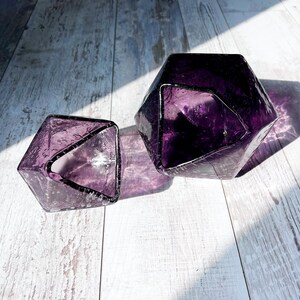 Puede incluir: Dos jarrones de cristal facetado de color amatista. Los jarrones tienen un dise&ntilde;o geom&eacute;trico con una superficie texturizada. La luz se refleja en el cristal, creando sombras. Los jarrones est&aacute;n sobre una superficie de madera clara.