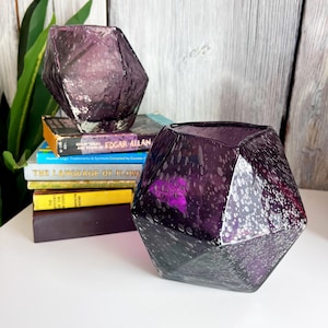 Puede incluir: Dos jarrones de cristal facetados de color amatista con un dise&ntilde;o moteado. Uno est&aacute; encima de una pila de libros, incluido uno titulado "EDGAR ALLAN". El otro jarr&oacute;n est&aacute; en primer plano, mostrando su forma geom&eacute;trica.