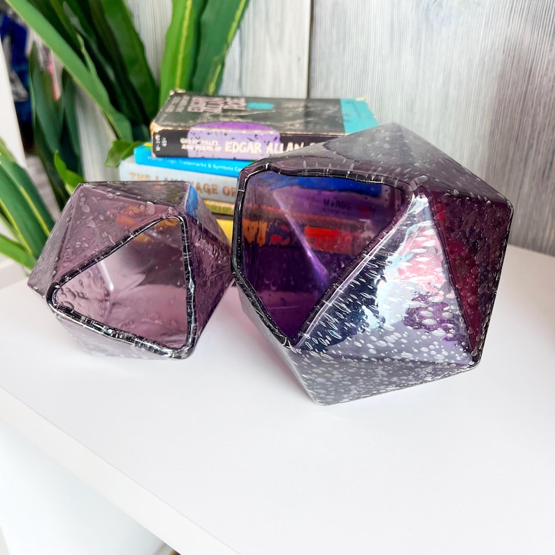 Puede incluir: Dos objetos geom&eacute;tricos de vidrio facetado en tonos p&uacute;rpura y amatista. Los objetos tienen un aspecto texturizado y moteado y est&aacute;n colocados sobre una superficie blanca. Los objetos son decorativos y podr&iacute;an usarse como jarrones o candelabros.