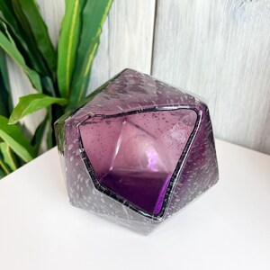 Puede incluir: Un jarr&oacute;n de cristal facetado de color amatista con un dise&ntilde;o geom&eacute;trico. El jarr&oacute;n tiene una abertura ancha y una superficie texturizada con peque&ntilde;as motas blancas. El interior es de un tono p&uacute;rpura m&aacute;s oscuro. El jarr&oacute;n est&aacute; sobre una superficie blanca.