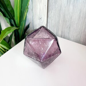 Puede incluir: Un jarr&oacute;n de cristal facetado de color amatista con un dise&ntilde;o geom&eacute;trico. El jarr&oacute;n tiene forma de diamante con superficies texturizadas y peque&ntilde;as burbujas de aire en el cristal. El jarr&oacute;n est&aacute; sobre una superficie blanca.