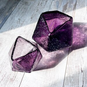 Puede incluir: Dos jarrones de cristal facetados de color amatista con una superficie texturizada y burbujeada. El dise&ntilde;o geom&eacute;trico de los jarrones crea sombras interesantes sobre la superficie de madera clara. Los jarrones est&aacute;n vac&iacute;os y parecen decorativos.