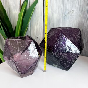Puede incluir: Dos jarrones de cristal facetados de color amatista. Los jarrones tienen un dise&ntilde;o geom&eacute;trico y una superficie texturizada. Un jarr&oacute;n mide aproximadamente 13 cm de alto. Los jarrones se exhiben sobre una superficie blanca con un fondo de madera.