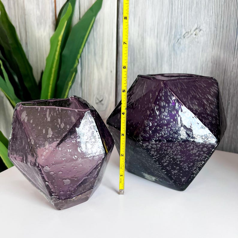 Puede incluir: Dos jarrones de cristal facetados de color amatista. Los jarrones tienen un dise&ntilde;o geom&eacute;trico y una superficie texturizada. Un jarr&oacute;n mide aproximadamente 13 cm de alto. Los jarrones se exhiben sobre una superficie blanca con un fondo de madera.