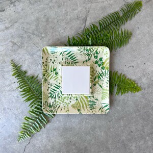 May include: A square decorative tray with a botanical design featuring green leaves and white flowers. A small white square is centered on the tray. Green fern fronds are arranged around the tray on a gray surface.