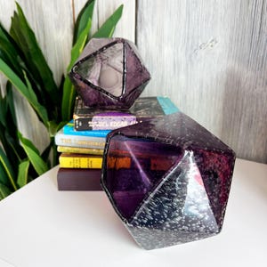 Puede incluir: Dos candelabros geom&eacute;tricos facetados de vidrio p&uacute;rpura. Los candelabros se colocan sobre una superficie blanca con una pila de libros en el fondo. El vidrio tiene una apariencia texturizada, similar a burbujas.