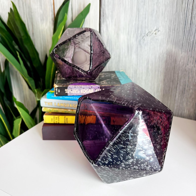Puede incluir: Dos candelabros geom&eacute;tricos facetados de vidrio p&uacute;rpura. Los candelabros se colocan sobre una superficie blanca con una pila de libros en el fondo. El vidrio tiene una apariencia texturizada, similar a burbujas.