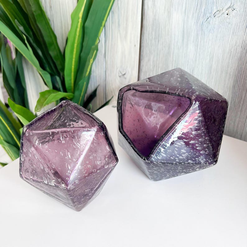 Puede incluir: Dos objetos decorativos de vidrio facetado de color amatista. Las formas geom&eacute;tricas tienen una superficie texturizada con peque&ntilde;as burbujas de aire. Los objetos se exhiben sobre una superficie blanca con follaje verde en el fondo.